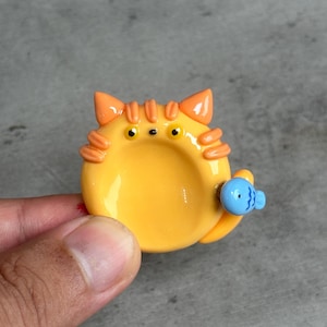 Könnte beinhalten: Eine kleine, runde, gelbe Keramikschale in Form eines Katzengesichts mit orangefarbenen gestreiften Ohren und Schnurrhaaren. Die Katze hat zwei gelbe Augen mit schwarzen Pupillen und eine winzige schwarze Nase. Ein kleiner, hellblauer Fisch mit schwarzen Liniendetails ist an der Seite befestigt.