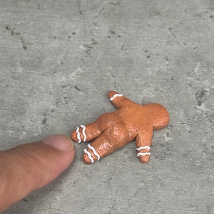 May include: A small, brown gingerbread person figurine lying face down. The figure has a textured surface and is decorated with thin, white, wavy icing lines around the wrists and ankles. The gingerbread person is positioned in a prone orientation.
