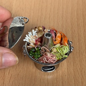 Puede incluir: Mini olla caliente plateada llena de comida colorida, incluyendo camarones, champiñones y fideos. La olla tiene una chimenea central y se encuentra sobre un pequeño soporte. Una tapa plateada se sostiene cerca.