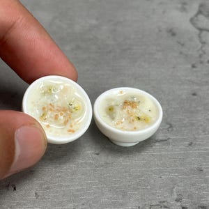 May include: Miniature white ceramic bowls filled with a creamy, off-white substance, speckled with green and brown bits, possibly representing a dessert or food item. The bowls are small, suggesting a dollhouse or craft project.