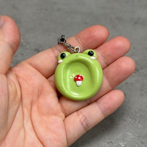 Può includere: Un ciondolo a forma di rana verde fatto a mano con occhi neri, guance rosa e un dettaglio a fungo rosso e bianco. Il ciondolo è attaccato a una catena e a una chiusura in metallo color argento. Il ciondolo a forma di rana è tenuto in una mano.