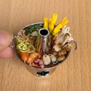 Pode incluir: Mini panela de prata cheia de uma variedade de alimentos, incluindo camarões, macarrão, cogumelos e flores amarelas. A panela tem uma chaminé central e pequenas alças. A panela é segurada por uma mão.