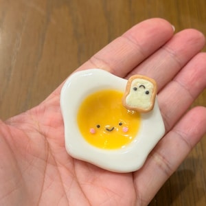 Puede incluir: Una pequeña figura de huevo frito, brillante, de color blanco y amarillo, con una cara sonriente y mejillas rosadas. Un trozo diminuto de pan tostado marrón claro con cara de sueño está unido al borde superior derecho del huevo, creando un adorno.