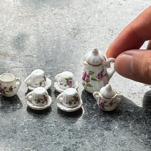 May include: Miniature porcelain tea set with a floral pattern in pink and purple. The set includes a teapot, creamer, sugar bowl, and four teacups with saucers. Gold trim accents the edges of the pieces.