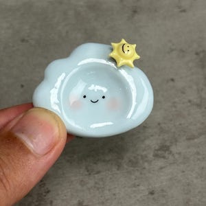 May include: A small, light blue ceramic cloud-shaped dish with a glossy finish. The cloud features a painted face with two black eyes, a small smile, and soft pink cheeks. A tiny, yellow, smiling sun figure is attached to the top right edge.