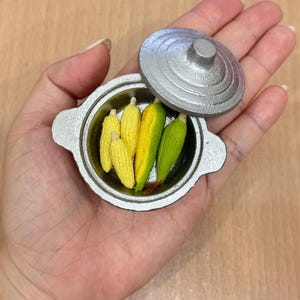 May include: Miniature silver pot with a lid, containing several small corn on the cob. The corn is yellow and green, with detailed husks. The pot has two handles and a round lid. The miniature food item is held in a hand.