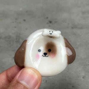 May include: A small, glossy ceramic dog figurine with a white face, brown ears, and a brown patch over one eye. The dog has pink cheeks, black eyes, and a small nose. A white bone-shaped piece with a black smiley face sits on top of its head.