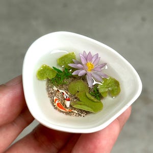 May include: A miniature white ceramic bowl containing a small pond scene with two tiny orange and white koi fish, a light purple water lily with a yellow center, several green lily pads, and small green aquatic plants set in clear resin.