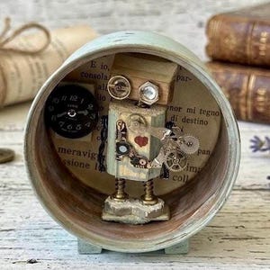Op de afbeelding: Een cirkelvormig lichtgroen frame met een kleine robotfiguur gemaakt van hout, metalen tandwielen en veren. De robot heeft een rood hart op de borst en staat voor een achtergrond van gedrukte Italiaanse tekst. Links is een zwarte klokplaat bevestigd.