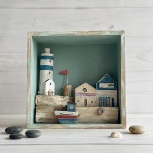 Puede incluir: Una caja de sombras de madera que representa una escena costera con un faro de rayas azules y blancas, casas y un barco. La escena incluye una bandera roja y la palabra "FISH". La caja está pintada en tonos azules y verdes.