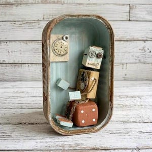 May include: A small robot sculpture constructed from wooden blocks, metal gears, and a clock face, housed inside a vertical oval metal tin. The robot sits on a brown cube base with white dots, featuring light blue limbs and a gear-adorned head.