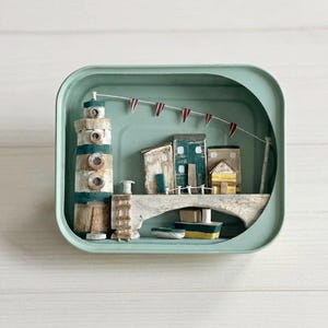 Könnte beinhalten: Ein Miniatur-Küsten-Diorama in einer türkisfarbenen Dose. Es zeigt einen Leuchtturm, kleine Gebäude, eine Brücke und ein Boot. Rote und weiße Wimpel hängen über der Szene. Das Kunstwerk ist aus Holz und in verschiedenen Farben bemalt.