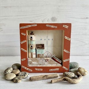 Puede incluir: Una caja de sombra de madera hecha a mano con una escena costera en miniatura. Incluye un faro, pequeñas casas y un barco, sobre un fondo de mapa. El marco es de color marrón rojizo, con etiquetas de texto. Guijarros y madera flotante están dispuestos delante.