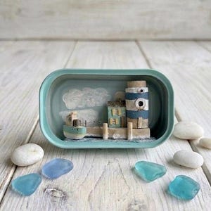 Könnte beinhalten: Miniatur-Küsten-Diorama in einem türkisfarbenen Behälter. Die Szene umfasst einen Leuchtturm mit blau-weißen Streifen, ein kleines Boot und ein Gebäude. Dekorative Meerglas- und glatte weiße Steine umgeben den Behälter und schaffen ein maritimes Thema.