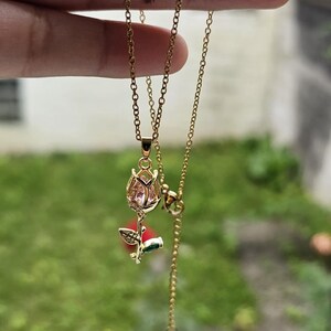 May include: Gold-colored necklace with a pendant featuring a rose bud design. The pendant has a pink gemstone center and a small butterfly charm. The chain is delicate and adjustable.