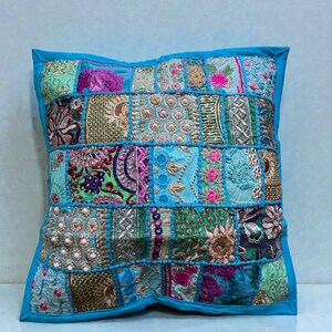May include: Square cushion with a patchwork design. The cushion features a variety of colourful fabric squares in shades of blue, green, pink, and gold, with intricate embroidery and beadwork. It has a solid blue border.