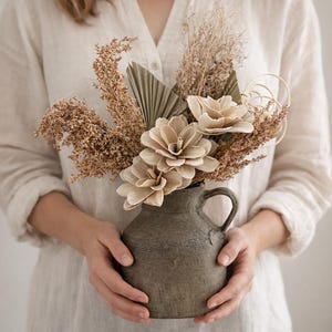 Puede incluir: Un jarrón de cerámica gris rústico con asa, lleno de flores secas y elementos decorativos. El arreglo incluye texturas beige, marrón claro y marrón, creando una estética natural y neutra. El jarrón es sostenido por una persona que lleva una camisa clara.