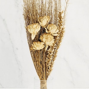 Op de afbeelding: Een droog bloemstuk met beige bloemen, tarwe en dunne bruine takken. Het boeket is vastgebonden met touw en wordt tentoongesteld in een heldere glazen vaas. De achtergrond is wit marmer.