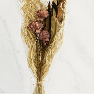 Puede incluir: Un ramo de flores secas en un jarrón de cristal transparente. El arreglo incluye tres flores de color malva, rodeadas de hojas secas y elementos similares a paja. El jarrón está atado con hilo, sobre un fondo de mármol blanco.