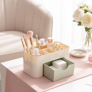 Op de afbeelding: Een beige make-up organizer met meerdere compartimenten en een uitschuifbare lade. De organizer bevat make-up borstels, parfumflesjes en andere schoonheidsproducten. De lade bevat watjes. De organizer staat op een roze ondergrond.