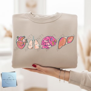 Peut inclure: Sweat-shirt beige avec une illustration de dessins animés d'organes humains : cœur, poumons, cerveau et foie. Les organes sont représentés en rose et rouge. Le sweat-shirt est tenu par une personne.