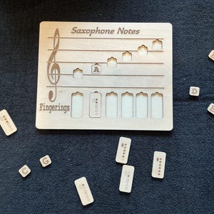 May include: A wooden saxophone fingering chart with the words "Saxophone Notes" and "Fingerings" engraved on it. The chart includes a treble clef, musical notes, and fingerings. Small wooden lettered blocks and fingering diagrams are scattered around the chart.