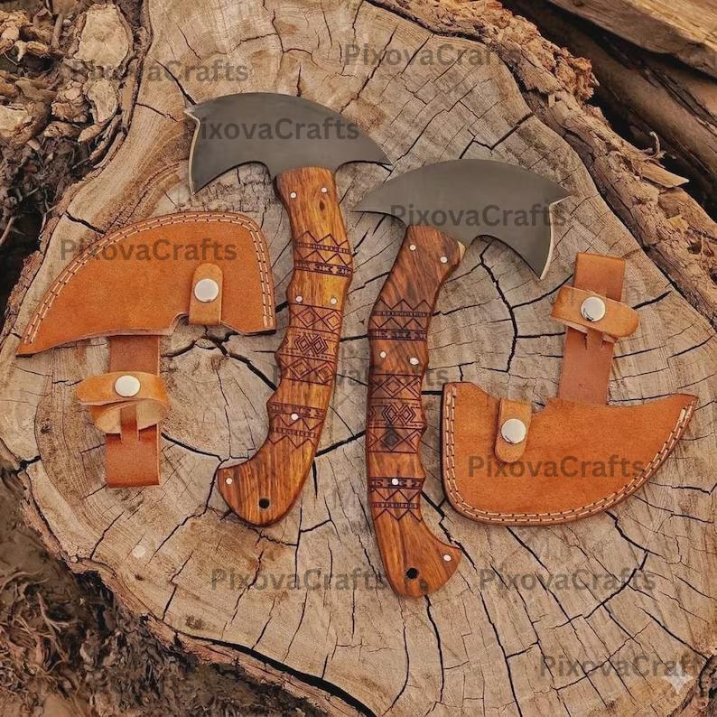 May include: Two curved blade axes with wooden handles and decorative carvings, accompanied by two leather sheaths. The axes and sheaths are displayed on a wooden surface. The handles are a light brown color, and the blades are a dark gray.