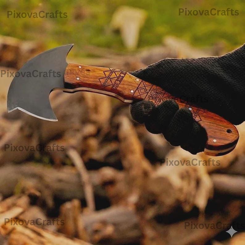 May include: A small hatchet with a dark blade and a wooden handle. The handle features decorative carvings and is held by a gloved hand. The hatchet is designed for outdoor use.