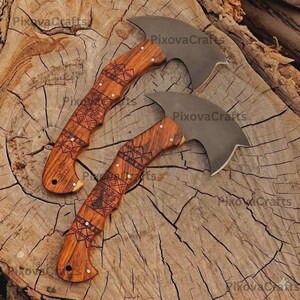 May include: Two small axes with curved blades and wooden handles. The handles are intricately carved with geometric patterns and accented with small metal studs. The blades are a dark gray color, and the wood has a warm, natural tone. The axes are displayed on a textured wooden surface.