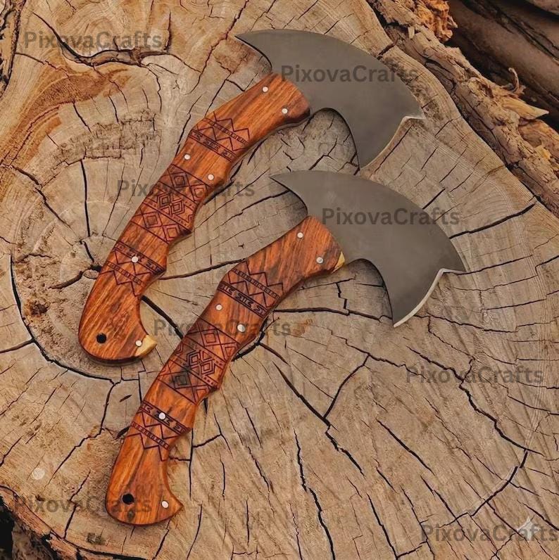 May include: Two small axes with curved blades and wooden handles. The handles are intricately carved with geometric patterns and accented with small metal studs. The blades are a dark gray color, and the wood has a warm, natural tone. The axes are displayed on a textured wooden surface.