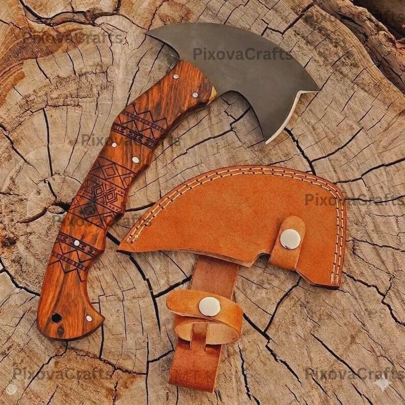May include: A handcrafted axe with a curved blade and a wooden handle featuring an engraved geometric design. The axe is accompanied by a brown leather sheath with a strap and button closure. The axe is displayed on a wood surface.