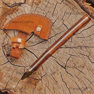 May include: A handcrafted hatchet with a wooden handle and a leather sheath. The hatchet has a brown blade and a silver stripe down the center of the handle. The leather sheath is tan and has a button closure. The text "PixovaCrafts" is visible.