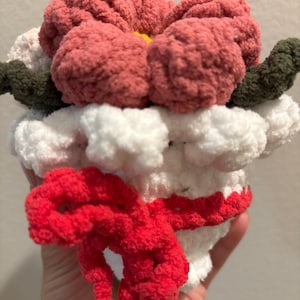 May include: A handmade crochet flower in shades of pink, white, and green. The flower has a fluffy texture and a red bow. The flower is a unique, handcrafted item.