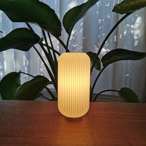 Puede incluir: Una lámpara de mesa acanalada de color crema brillante se encuentra sobre una superficie de madera. La lámpara tiene forma cilíndrica con ranuras verticales. Hojas verdes de plantas en el fondo, con una cortina blanca transparente visible.