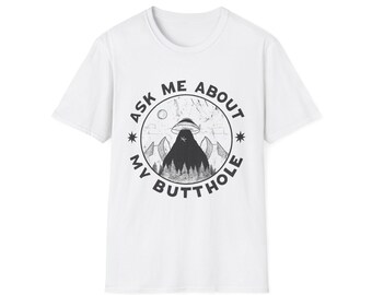 May include: White t-shirt with a black and white graphic. The graphic features a mountain scene with a UFO and the text "ASK ME ABOUT MY BUTTHOLE" in a circular design. The shirt has a crew neck and short sleeves.