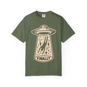 Op de afbeelding: Olijfgroen t-shirt met een beige afbeelding van een UFO die een persoon ontvoert. Het ontwerp bevat sterren, kruisen en het woord "FINALLY". Het shirt heeft korte mouwen en een ronde hals.
