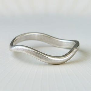 Può includere: Un anello color argento con un design ondulato. L'anello è realizzato in metallo liscio e lucido e si trova su una superficie bianca. La forma dell'anello ricorda una dolce onda.