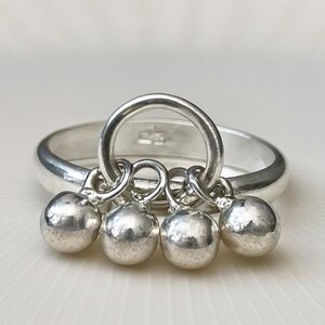 May include: A silver ring with a circular design and four dangling silver spheres. The ring is set against a white background, highlighting the polished metal and the ring's unique design. The spheres are evenly spaced and add a playful element to the jewelry.