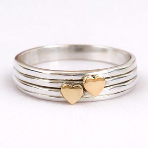 Puede incluir: Un anillo de plata con tres bandas y dos pequeños acentos de corazón dorado. El anillo está sobre una superficie blanca. El anillo es una joya.