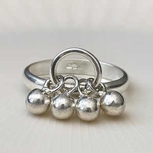 May include: A silver ring with a circular design and four small, dangling spheres. The ring is set against a white background, highlighting its polished, reflective surface. The spheres are evenly spaced and add a playful element to the jewelry.