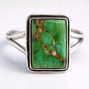 Peut inclure: Bague en argent ornée d'une pierre rectangulaire vert vif veinée de brun. La pierre est sertie dans un cadre en argent, et la bague a un design à tige fendue. La bague est présentée sur une surface blanche.