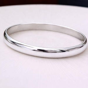 May include: A polished silver bangle bracelet. The bracelet has a smooth, rounded design and reflects light, creating a bright, shiny appearance. The jewelry is a simple, elegant accessory.
