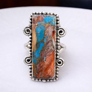 Könnte beinhalten: Silberner Ring mit einem rechteckigen Stein. Der Stein zeigt eine Mischung aus Türkis, Braun und Gold. Der Ring hat einen dekorativen Rand mit kleinen Perlen und scrollartigen Verzierungen an den Seiten. Ein Schmuckstück.