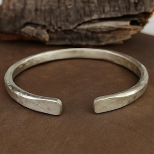 Könnte beinhalten: Ein silbernes Armreif-Armband mit gehämmertem Muster. Das offene Design hat flache, rechteckige Enden. Das Armband wird auf einer braunen Oberfläche mit einem Stück Treibholz im Hintergrund präsentiert.