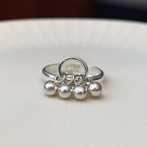 May include: A silver ring with a circular design and four small, dangling spheres. The ring is set against a white background, highlighting its polished, reflective surface. The spheres are evenly spaced and add a playful element to the jewelry.