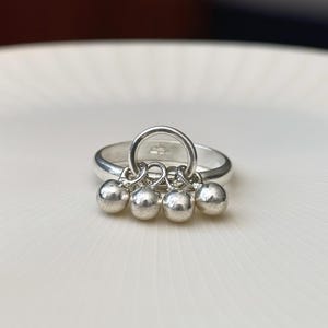 May include: A silver ring with a circular design and four small, dangling spheres. The ring is set against a white background, highlighting its polished, reflective surface. The spheres are evenly spaced and add a playful element to the jewelry.