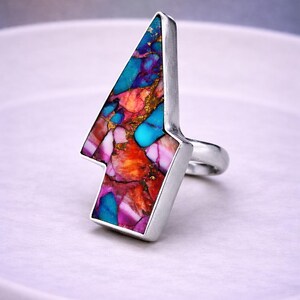 Op de afbeelding: Een zilveren ring met een pijl-vormige inleg bestaande uit veelkleurige steenfragmenten in tinten turquoise, magenta, oranje en paars, geaccentueerd met metallic gouden spikkels in een frame en band van gepolijst zilver.