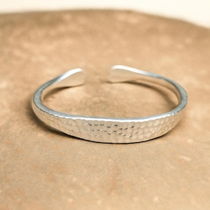 May include: A silver cuff bracelet with a hammered finish. The open design features a slightly rounded band with a textured surface. The bracelet is displayed on a neutral background.