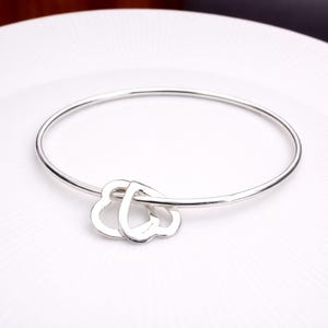 Könnte beinhalten: Ein silbernes Armreif-Armband mit einem doppelten Herzanhänger. Das Armband besteht aus dünnem, poliertem Metall und hat zwei herzförmige Anhänger, die sich in der Mitte verschlingen. Das Armband wird auf einer weißen Oberfläche präsentiert.
