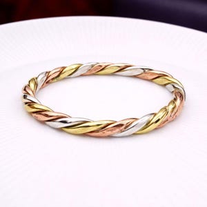Può includere: Un braccialetto intrecciato tricolore. Il braccialetto presenta fili intrecciati d'oro, argento e oro rosa, creando un accessorio strutturato e accattivante. Il braccialetto è circolare e si trova su una superficie bianca.